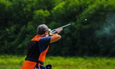 A shooter in an orange vest aims a shotgun at a clay target against a green backdrop, illustrating the sport of clay pigeon shooting.