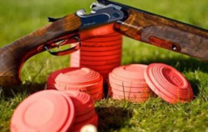 A shotgun rests on green grass beside stacked red clay targets, showcasing a clay shooting setup for sports enthusiasts.