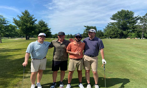 Four men smiling together in front of a golf cart parked on a paved path.