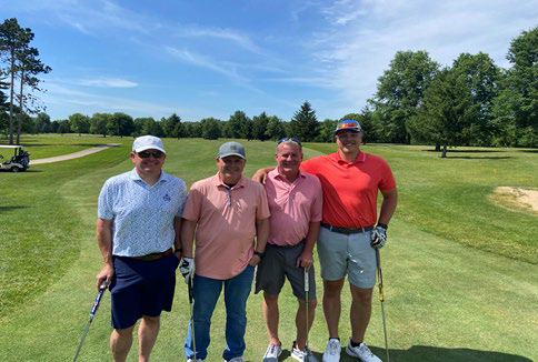Four men in golf polo shirts and hats posing together on a grassy area.