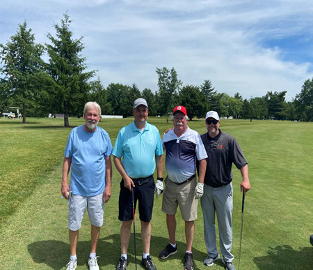 Four men posing together on a golf course pathway under a slightly cloudy sky.