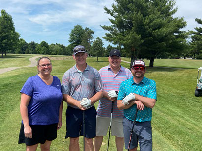 Four men smiling together next to a white golf cart on the course pathway.