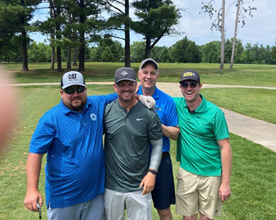 Four men in golf shirts and caps hugging and laughing for a group photo.
