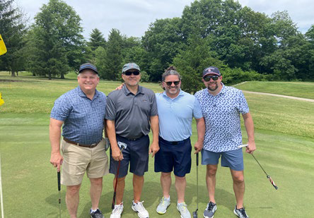 Four men smiling together on a putting green under an overcast sky.
