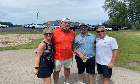 A woman and three men posing near a parking lot, with two of the men holding cigars.
