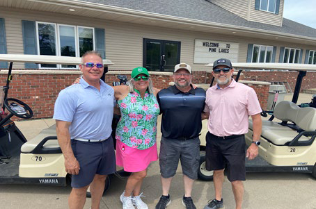 Three men and a woman in a vibrant floral outfit posing in front of the Pine Lakes clubhouse with golf carts.