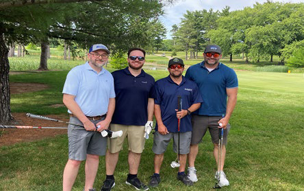 Four men in polo shirts holding golf clubs under a shade tree on the course.