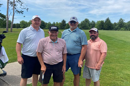 Four men smiling together on a golf course, with one wearing a hat that reads "CUPP".