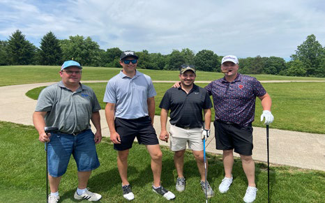 Four men in golf attire posing on a green fairway, one holding a cigar.