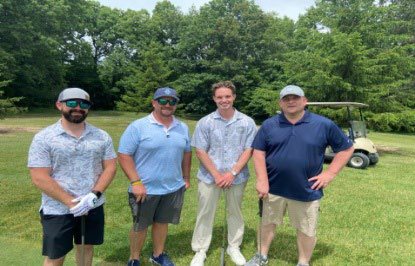 Four men in golf attire standing next to a golf cart on a paved path.