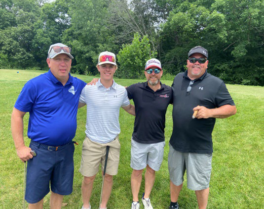 Four men in athletic shirts smiling and posing together on a grassy lawn.