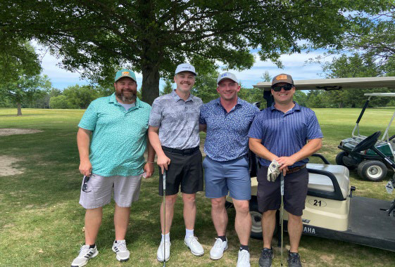 Four men in golf clothes standing in a row on the grass next to a paved path.
